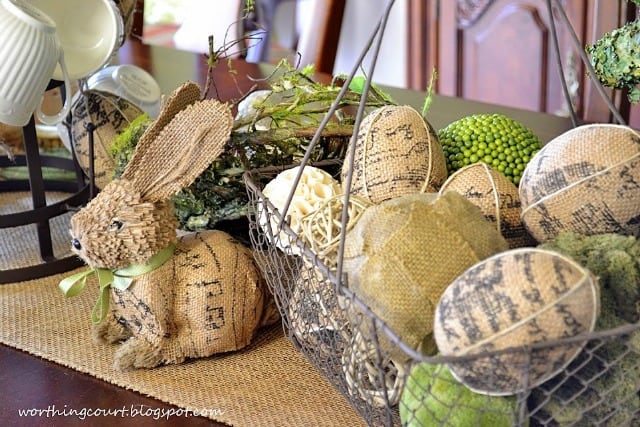 An Easter centerpiece with an aged wire basket filled with burlap eggs and a variety of decorative orbs flanked by a burlap bunny.