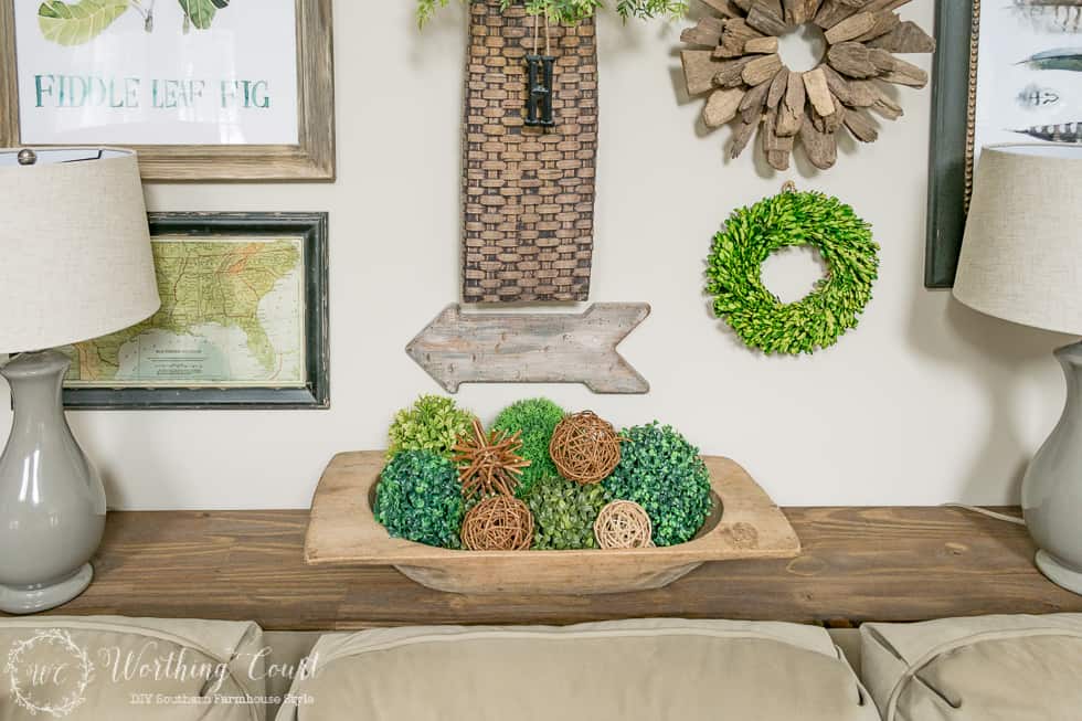 There is a wooden dough bowl on the table filled with greenery and twig balls.