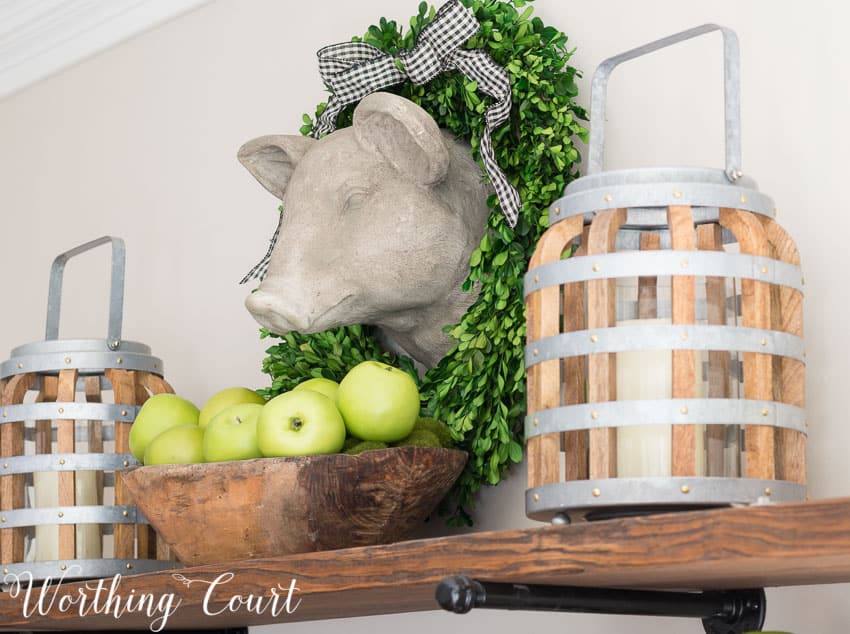 A faux pig head, surrounded by a preserved boxwood wreath, hangs above open rustic farmhouse kitchen shelves