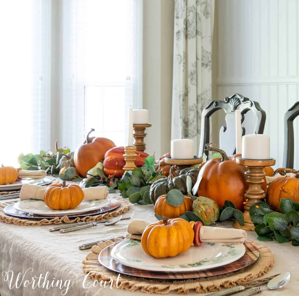 a Thanksgiving centerpiece using various sizes of faux pumpkins on a bed of eucalyptus leaves with wooden candle holders