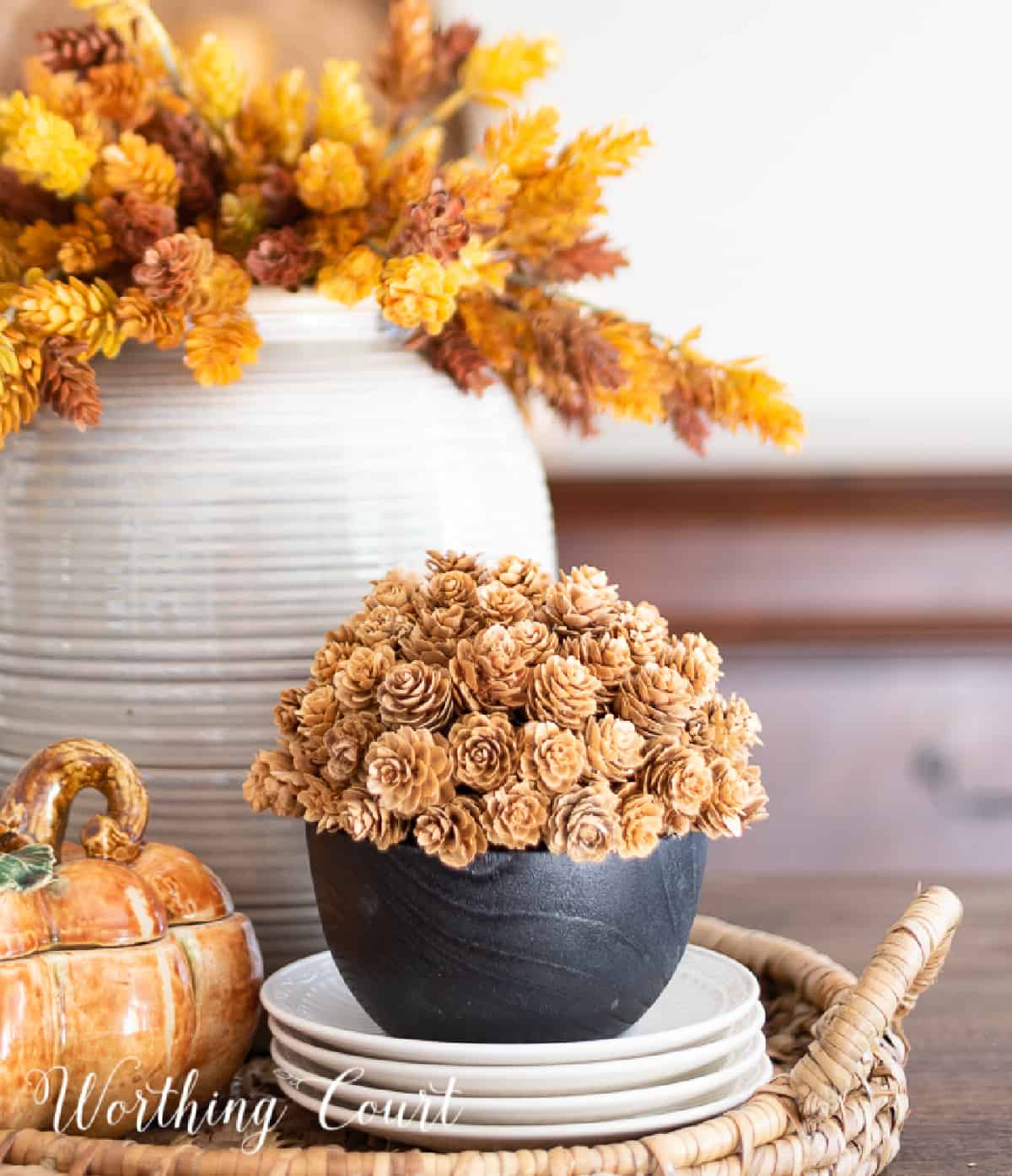 Thanksgiving centerpiece with a vase of fall stems, porcelain pumpkin box and small pinecone arrangement in a round wicker tray