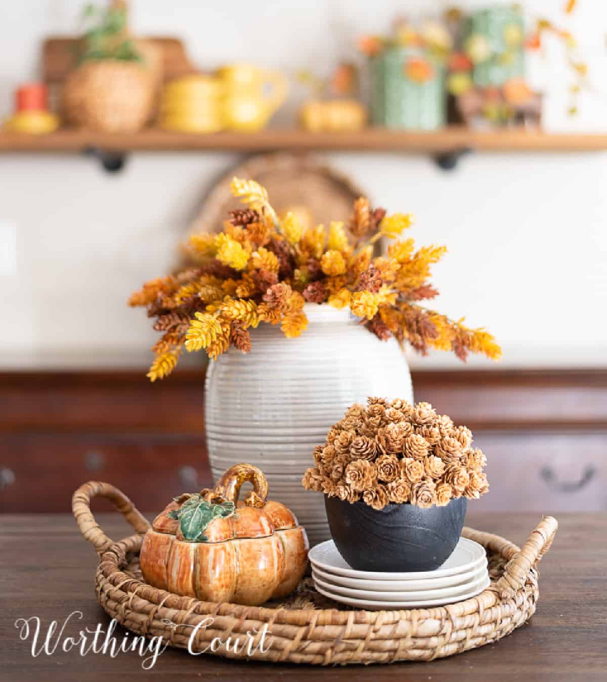 Thanksgiving centerpiece with a vase of fall stems, porcelain pumpkin box and small pinecone arrangement in a round wicker tray