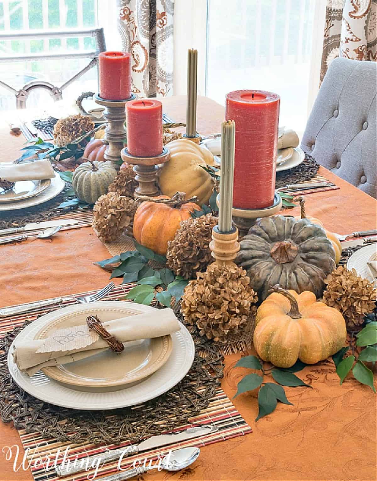 Thanksgiving centerpiece with orange and green candles surrounded by a bed of foliage and dried hydrangeas with faux pumpkins