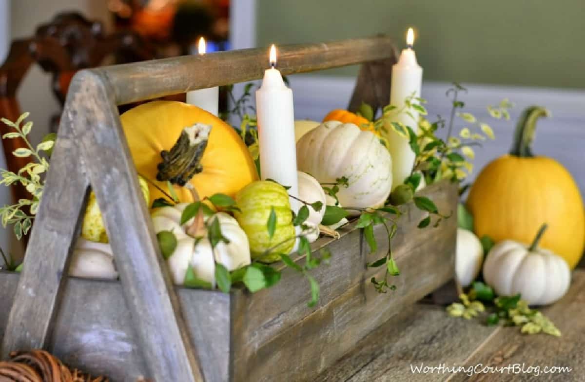 long gray wood tool box filled with yellow and white pumpkins, white candles and sprigs of greenery for a Thanksgiving centerpiece