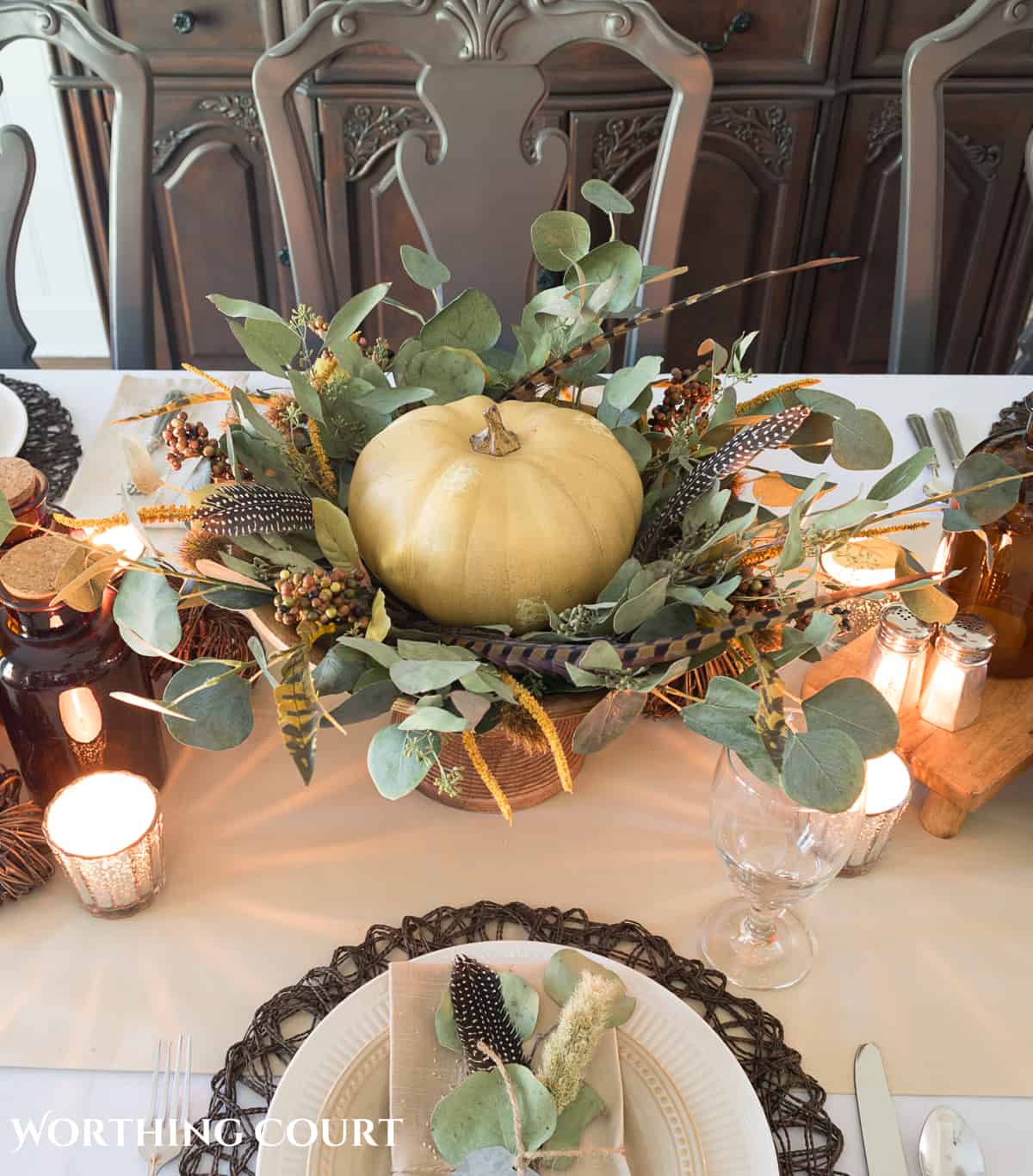 Thanksgiving centerpiece with cream colored pumpkin on a wooden pedestal surrounded by bits of greenery and feathers