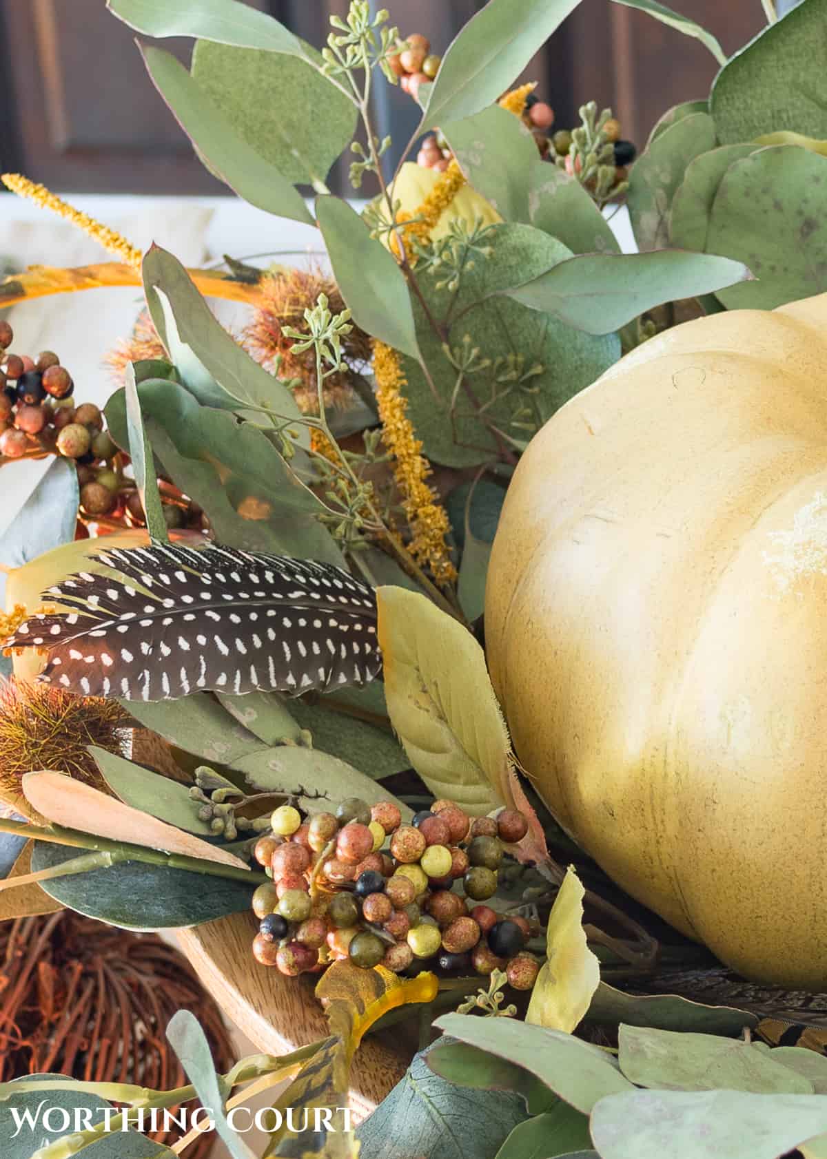 Thanksgiving centerpiece with cream colored pumpkin on a wooden pedestal surrounded by bits of greenery and feathers