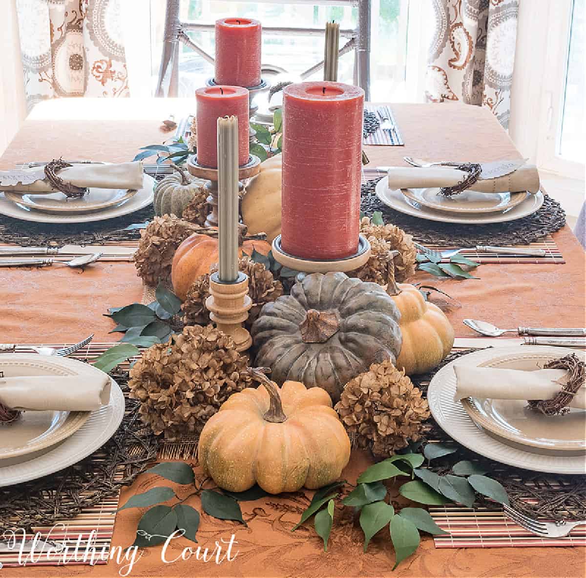 Thanksgiving centerpiece with orange and green candles surrounded by a bed of foliage and dried hydrangeas with faux pumpkins