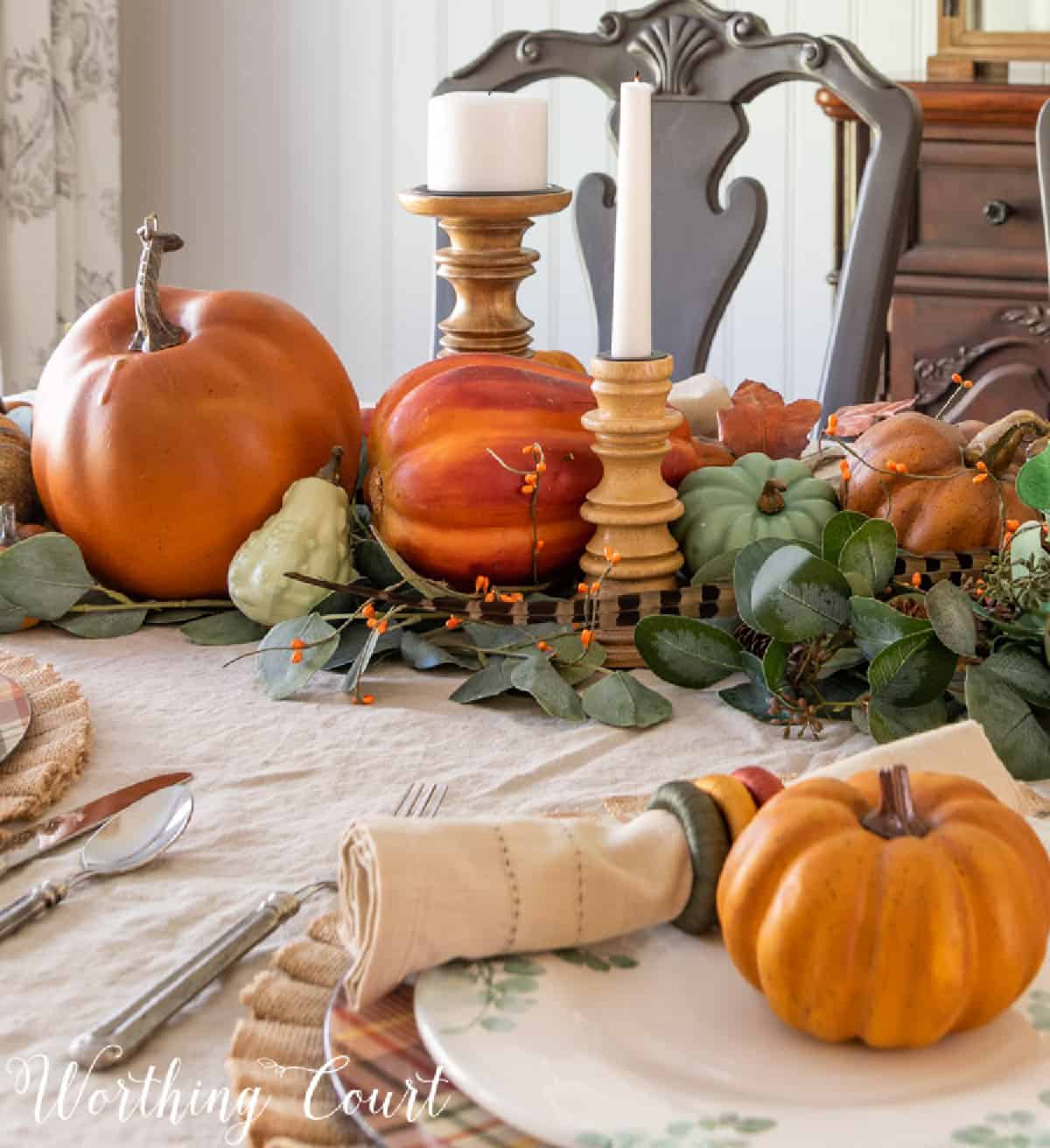 a Thanksgiving centerpiece using various sizes of faux pumpkins on a bed of eucalyptus leaves with wooden candle holders