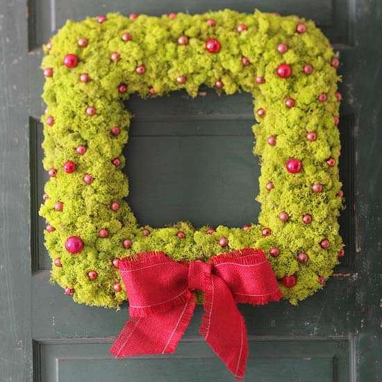 A square wreath in bright green and pinkish/red details with a red bow hanging on a door.