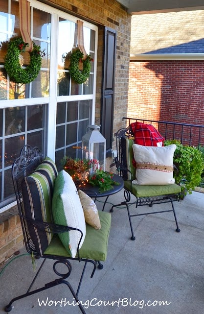Two wrought iron chairs with green pillows and a tartan blanket. There are small wreaths on the outdoor windows.