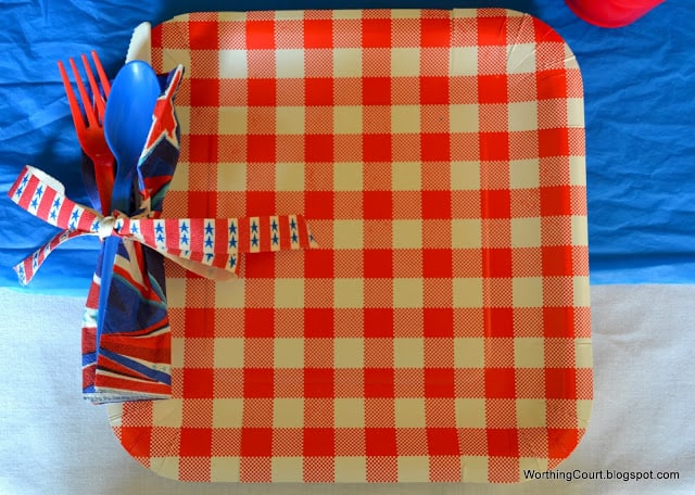 Super łatwe patriotyczne ustawienie miejsca za pomocą papierowych talerzy, papierowych serwetek i plastikowych przyborów, wszystko ze sklepu dollar. Użyj dziurkacza, aby wbić otwór w bok płyty. Przeprowadź wstążkę przez otwór i zawiąż ją wokół zwiniętych papierowych serwetek i plastikowych przyborów.