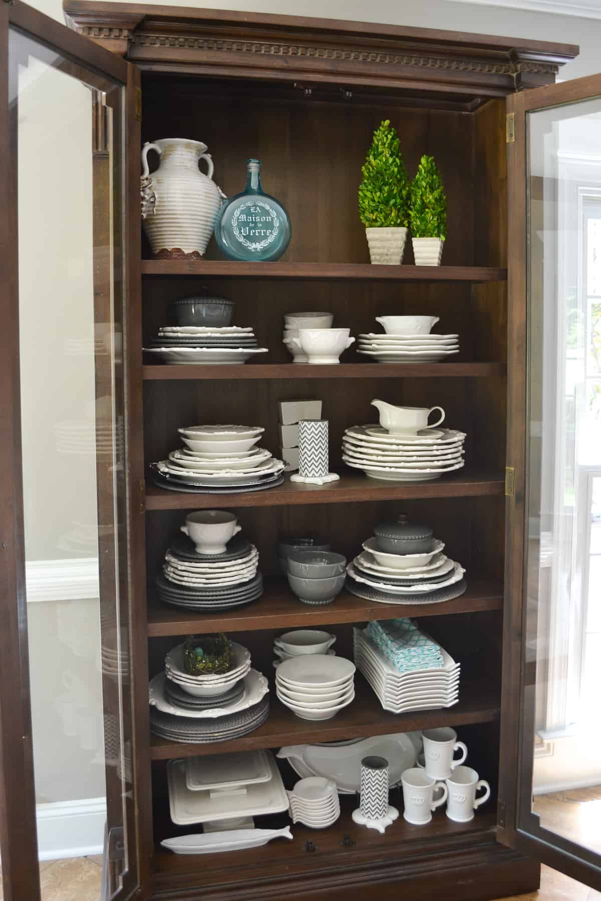 tall wood cabinet with glass doors filled with white plates, platters and bowls