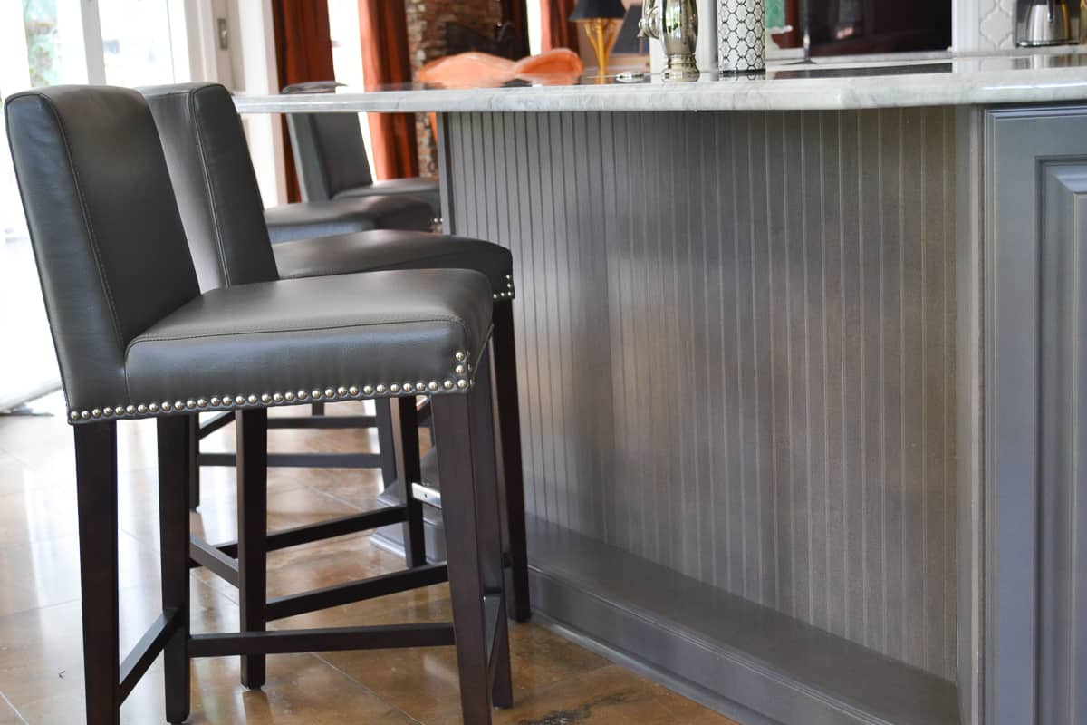 front of gray kitchen island covered with beadboard paneling and gray leather barstools