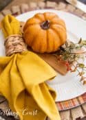 fall place setting with a burlap placemat, fall plaid charger and white plate topped with fall decor and a yellow napkin