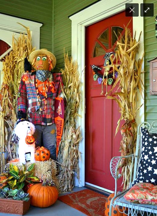 A small porch with a red door and the Halloween decor has a scarecrow, pumpkins and wheat stalks .