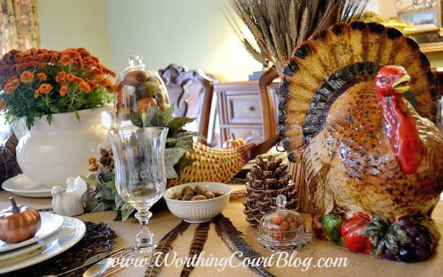 Thanksgiving tablescape with a large porcelain turkey, flowers, wine glasses and pine cones on the table.