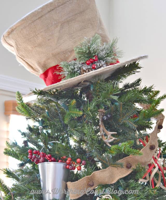 Burlap snowman hat Christmas tree topper with holly and a ribbon around it.