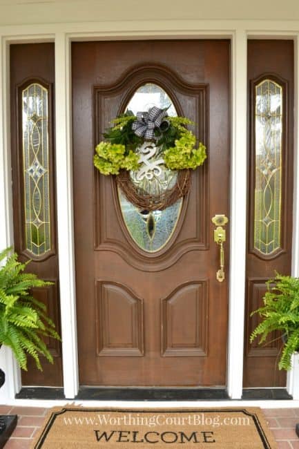 The spring wreath hanging on the door with a welcome mat outside of it.