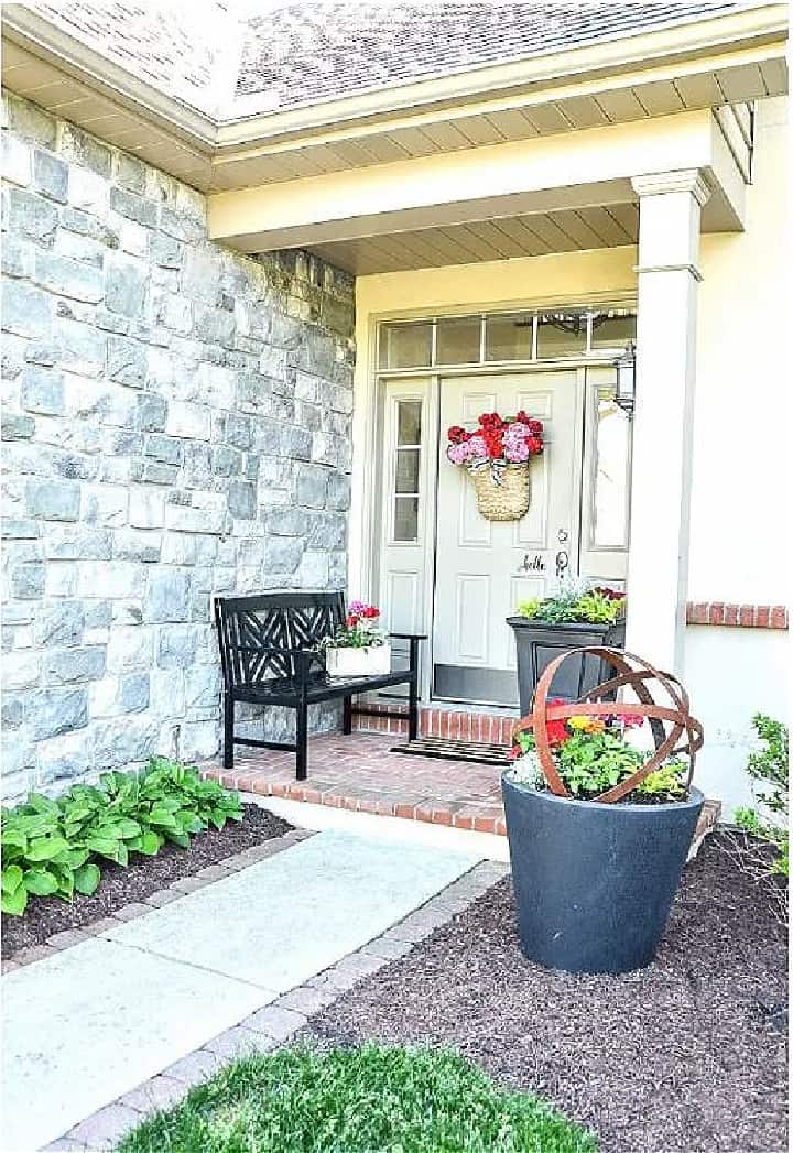 front porch on a stone house with a basket of flowers on the door and a black bench