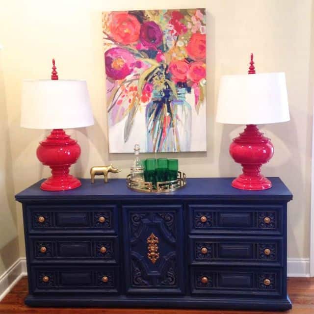 There is a blue sideboard with red lamps and a colorful picture on the wall.