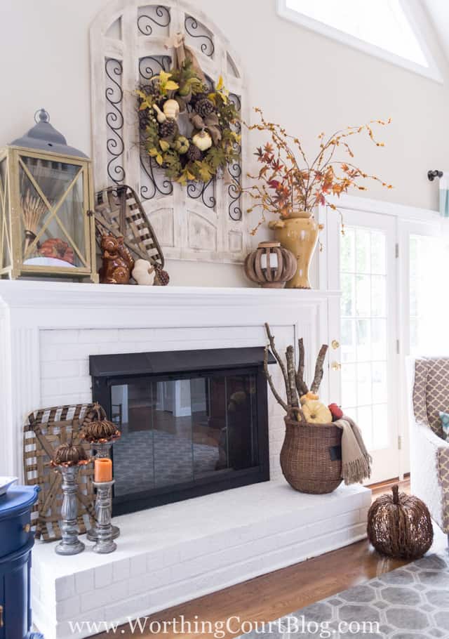 A fall wreath is hanging above the fireplace mantel, with fall decor items on the white fireplace mantel.   There is a wicker basket below beside the fireplace.