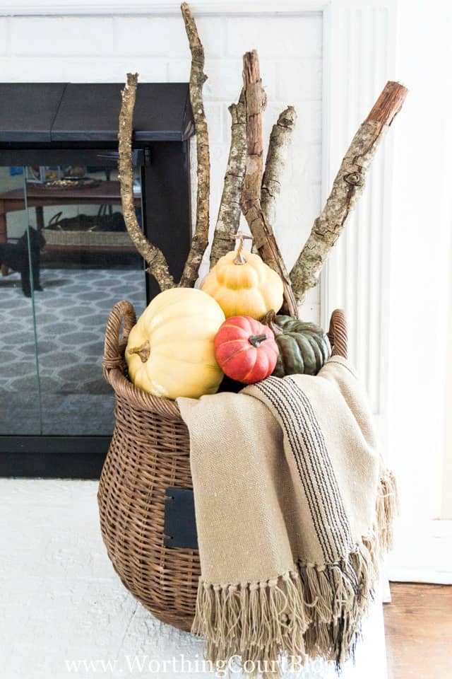 Sticks, branches, small pumpkins and a throw blanket are in the wicker basket beside the fireplace.