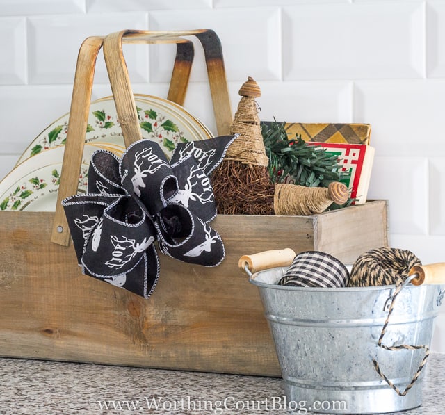 A wooden carrying box filled with kitchen plates, ribbon and mini Christmas trees.