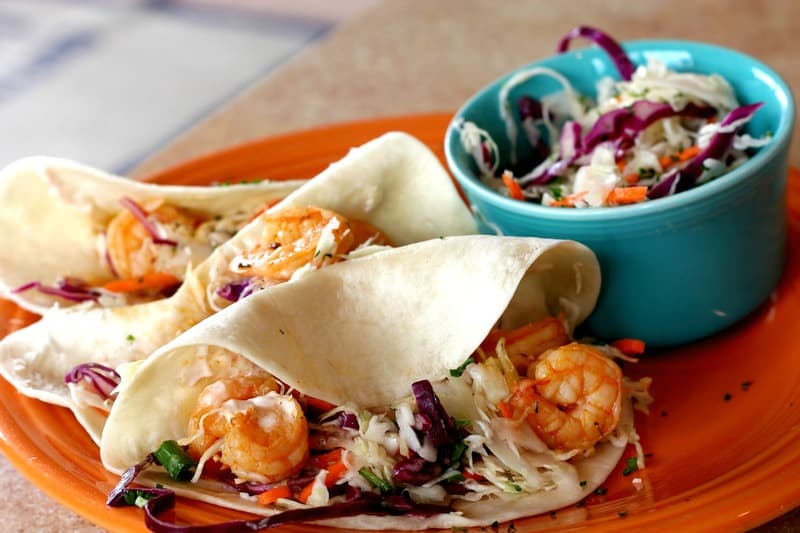 The shrimp tacos on a orange plate with a side of slaw in a blue bowl.