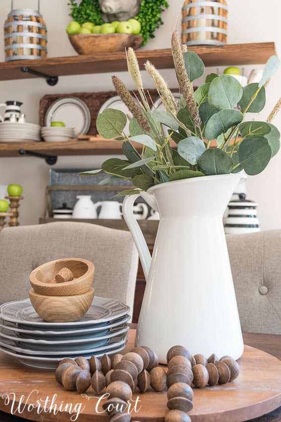 A white pitcher and dishes with wooden beads all on a cutting board in the dining room.