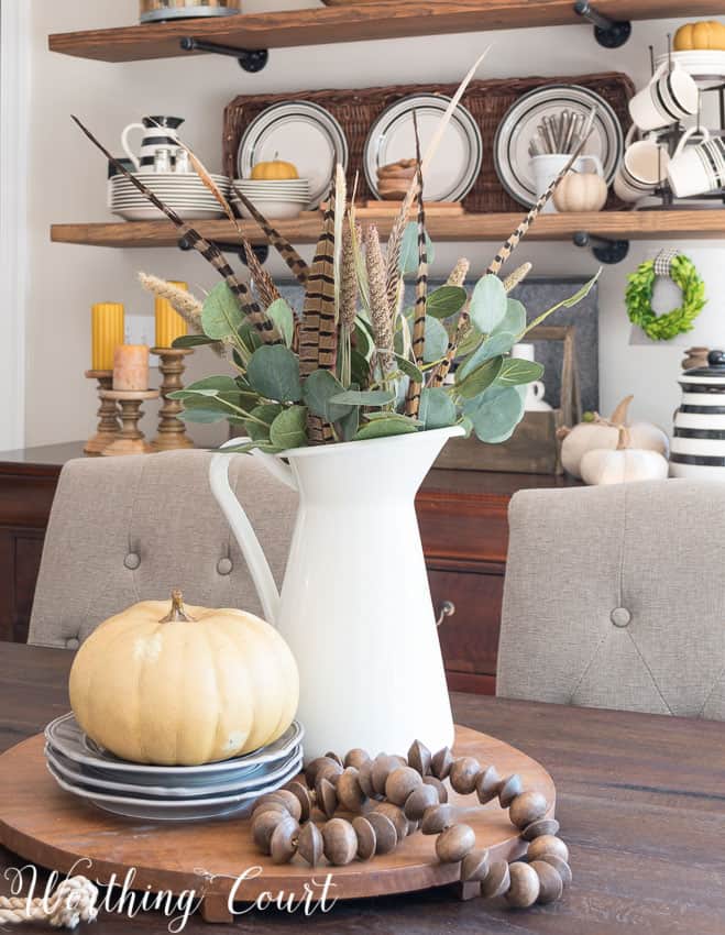 A white pitcher is filled with eucalyptus leaves, feathers and it is on a wooden tray that is round on the table.