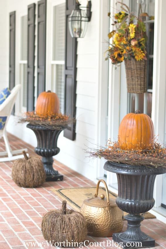 Two large urns with twig wreaths and pumpkins on top of the wreaths all on the front porch.