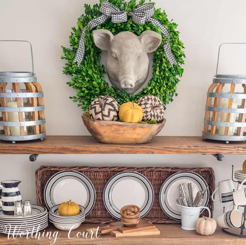 Herbert the pig has a wreath with a checkered bow around him and a dough bowl filled with fall items underneath on the shelves.