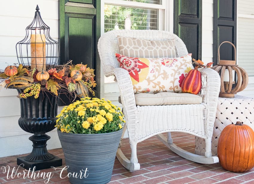 Farmhouse style fall porch vignette with a white wicker rocking chair, pumpkins and a large urn with a candle in it.