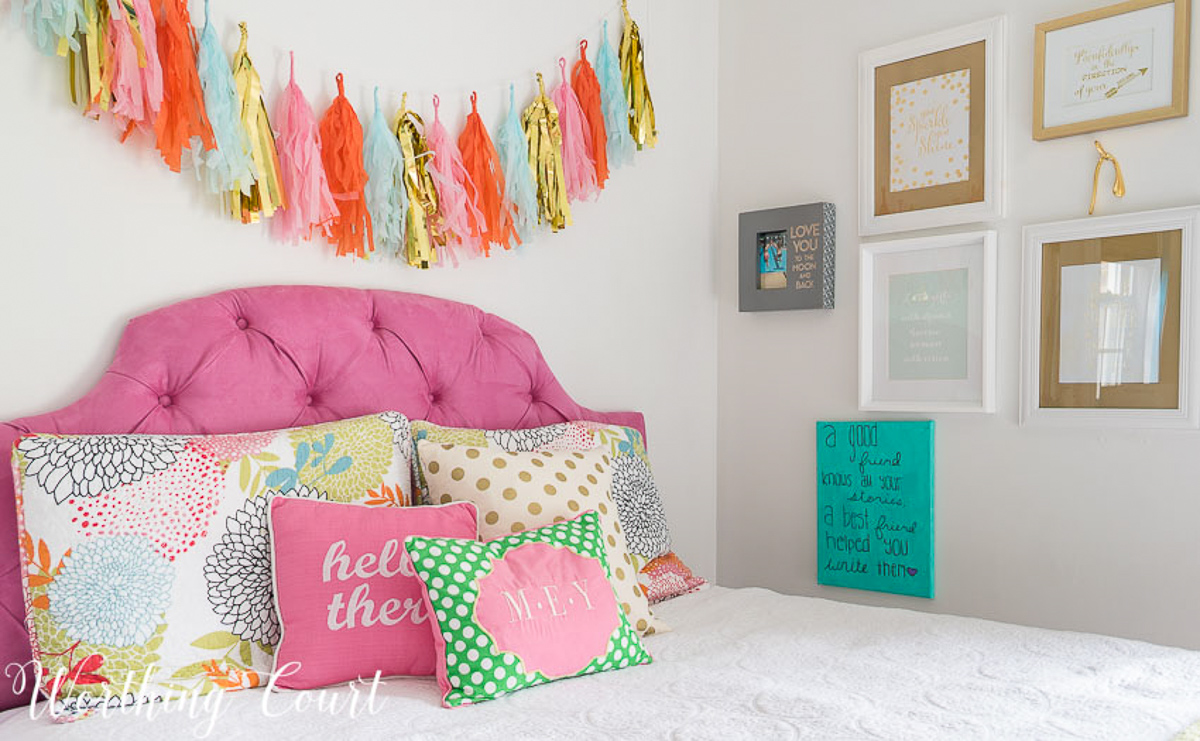 colorful teenage girls bedroom with a pink headboard, furniture painted white, teal lamp shades, teal throw, brass trunk, tissue paper garland and a gallery wall
