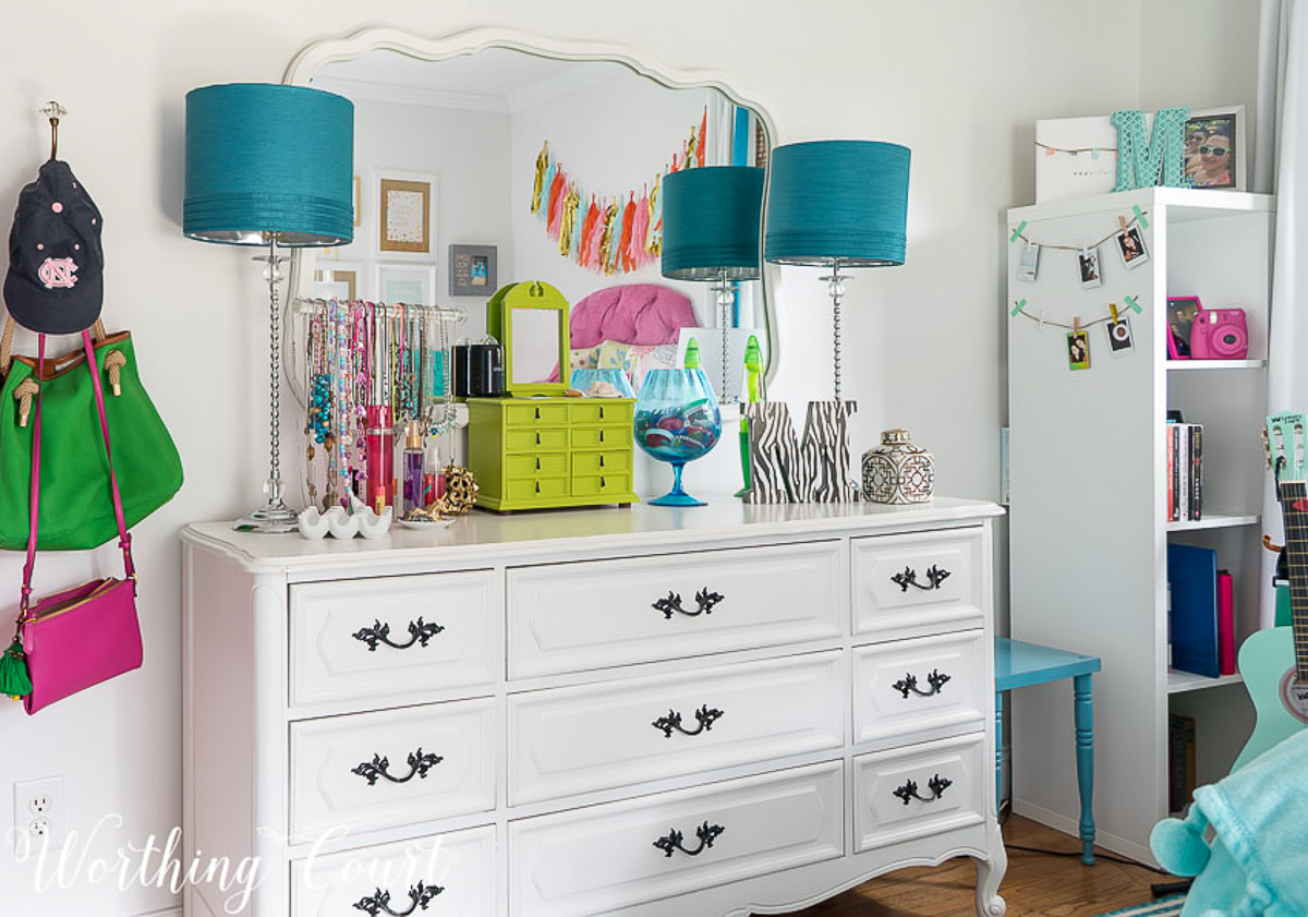 colorful teenage girls bedroom with a pink headboard, furniture painted white, teal lamp shades, teal throw, brass trunk, tissue paper garland and a gallery wall