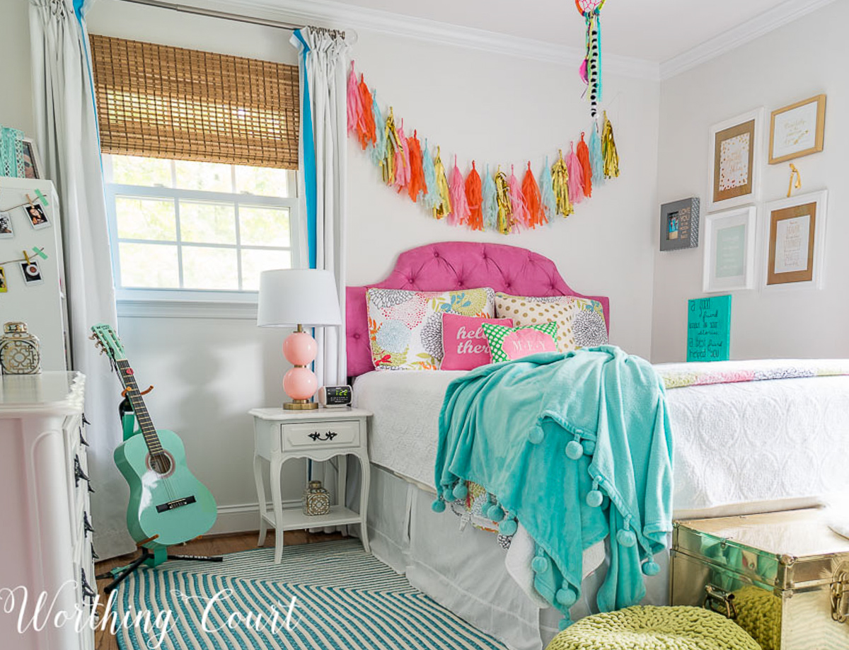 colorful teenage girls bedroom with a pink headboard, furniture painted white, teal lamp shades, teal throw, brass trunk, tissue paper garland and a gallery wall