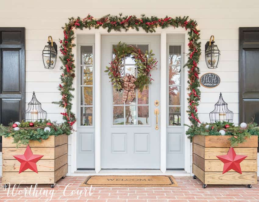 Front door surrounded with lighted garland and flanked with diy planters for Christmas.