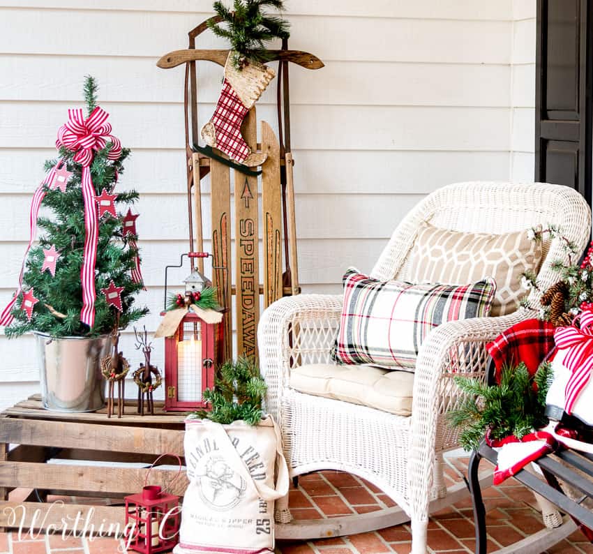 A porch decorated with a mini Christmas tree, a white rocking chair with a buffalo checked pillow and a wooden sled.