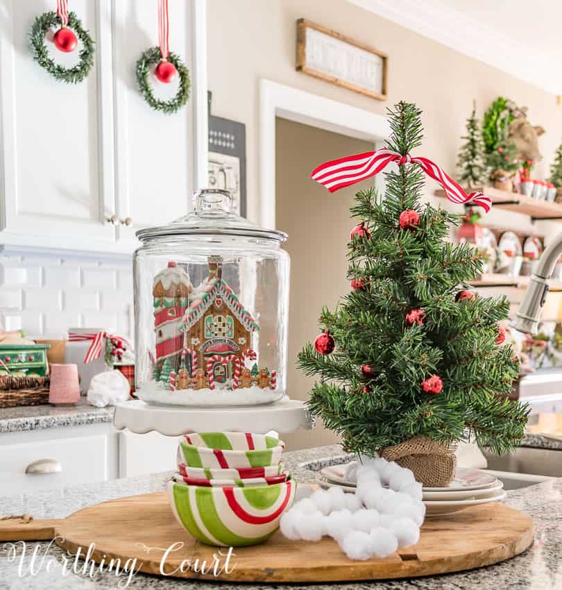 Christmas kitchen island vignette featuring a Christmas village house in a glass canister and a mini decorated Christmas tree on the counter.