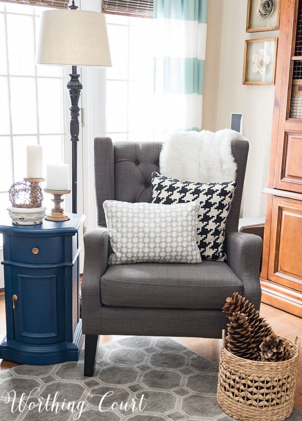 A gray armchair with throw pillows on it, and a woven basket full of pinecones.