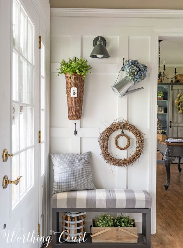 A decorated board and batten wall with a small wreath, watering can with flowers, and a wicker basket with greenery in it.