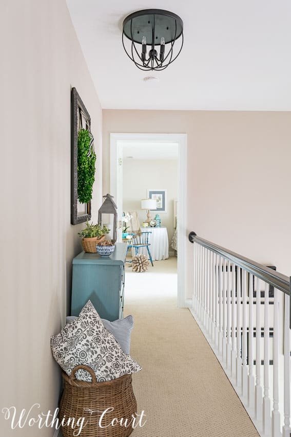 There is woven basket filled with throw pillows beside the chest of drawers in the hallway.