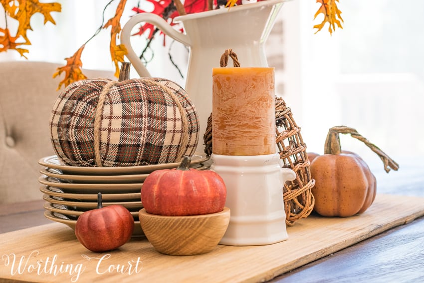 A plaid pumpkin, candles and a white pitcher filled with fall leaves.