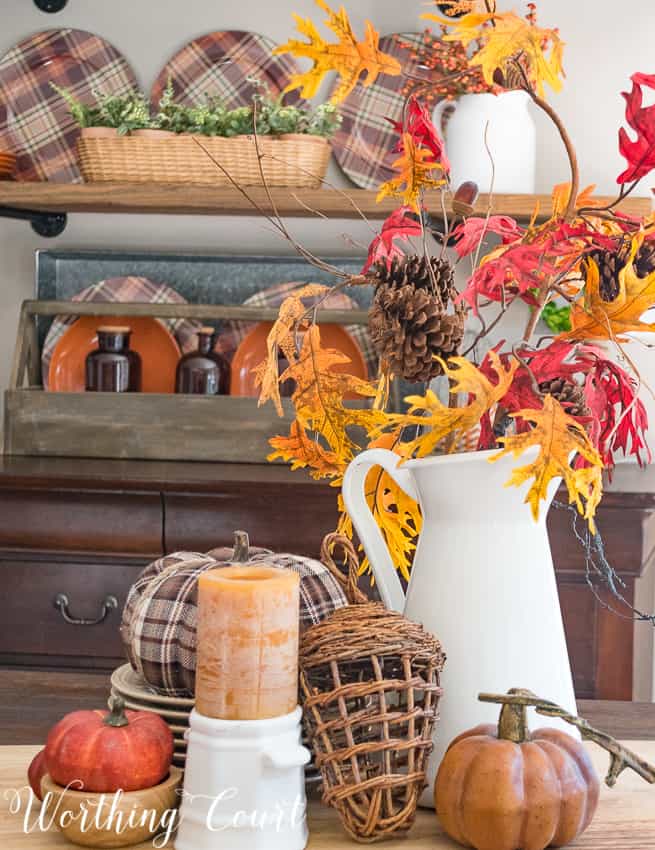 A white pitcher on the table filled with fall leaves, a plaid pumpkin, a candle and small pumpkins.