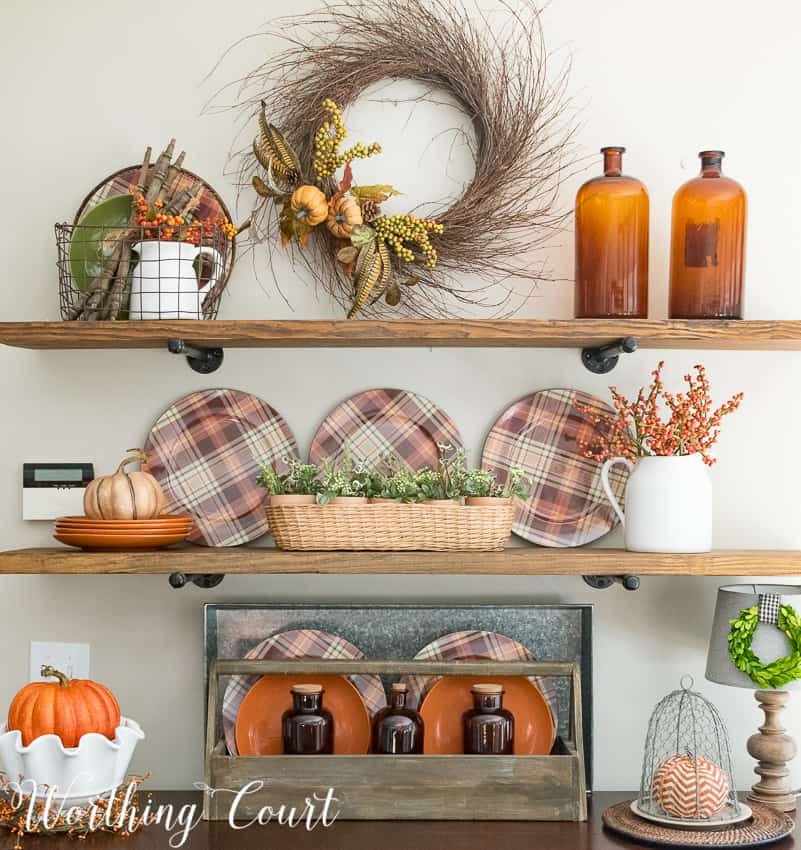 Open shelving with a fall wreath on the wall, plaid plates, and amber glass.