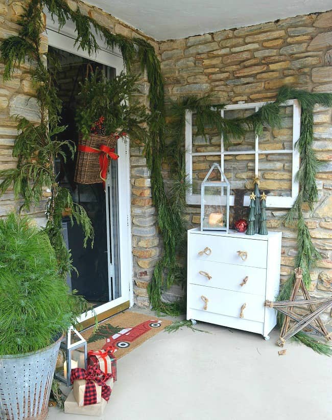 The front porch of a house with a brick facade and a basket hanging on the door filled with evergreen.