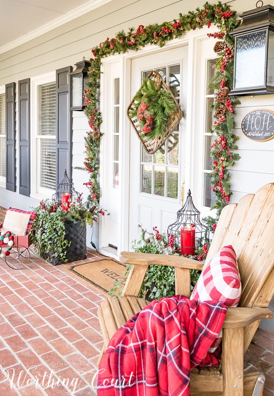 The front porch decorated with an adirondack chair with a red and white pillow and a throw blanket.