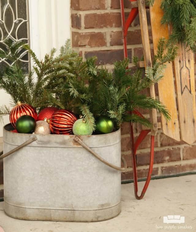 A galvanized bucket is filled with ornaments and evergreen branches on the porch.