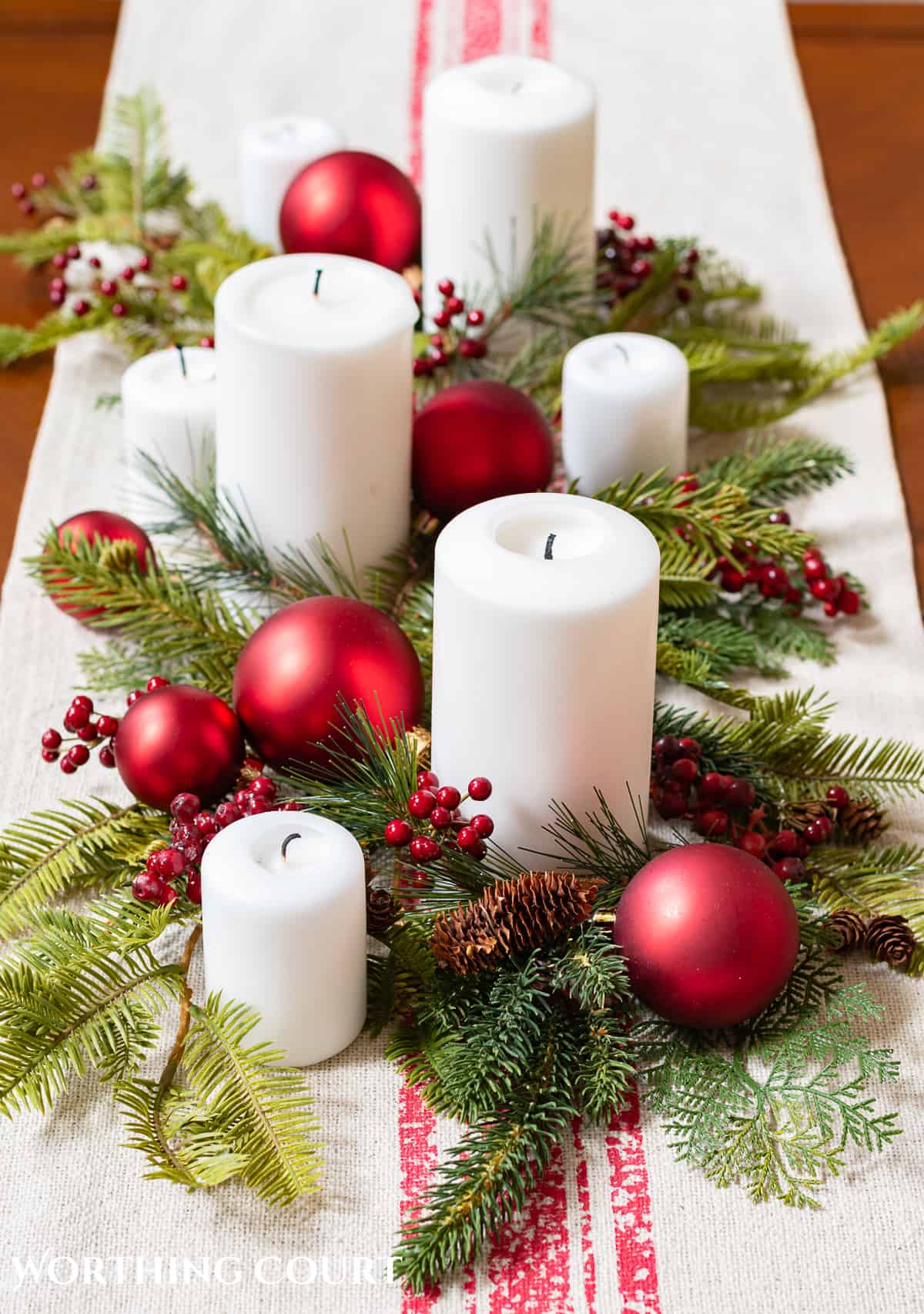 Assorted white candles on a coffee table with faux greenery and red Christmas ornaments on top of a table runner