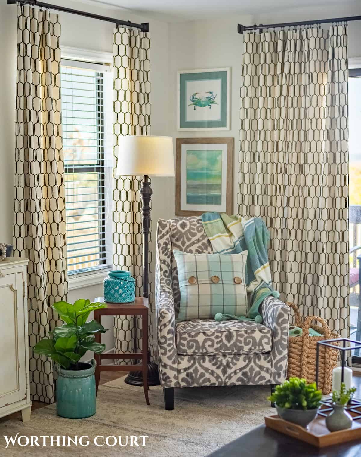 gray and white arm chair in corner in front of windows with neutral curtains with lining and with blue and aqua pillow and coastal art above